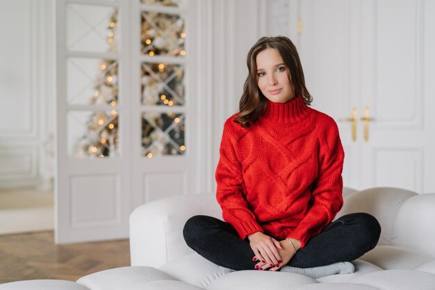 Hausfrau in rotem Pullover sitzt in Lotus-Pose auf der Couch Neujahrsbaum im Hintergrund Ruhe-Konzept