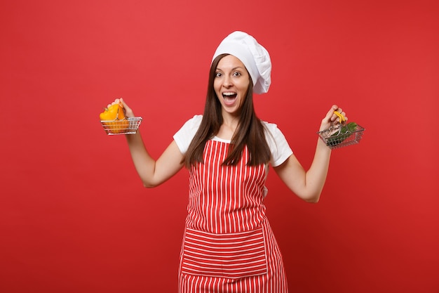 Hausfrau Köchin Koch Bäcker in gestreifter Schürze weißes T-Shirt Haube Kochmütze isoliert auf rotem Wandhintergrund. Frau hält gelbe Paprika-grüne Gurke im Einkaufswagen Mock-up-Kopierraumkonzept
