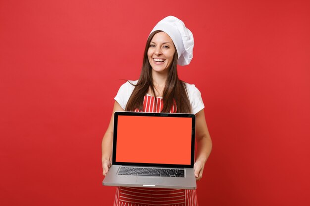 Hausfrau Köchin oder Bäckerin in gestreifter Schürze weißes T-Shirt Haube Kochmütze isoliert auf rotem Wandhintergrund. Frau hält Laptop-PC-Leerbildschirm für Werbeinhalte Mock-up-Kopierraumkonzept