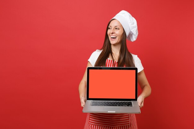 Hausfrau Köchin oder Bäckerin in gestreifter Schürze weißes T-Shirt Haube Kochmütze isoliert auf rotem Wandhintergrund. Frau hält Laptop-PC-Leerbildschirm für Werbeinhalte Mock-up-Kopierraumkonzept