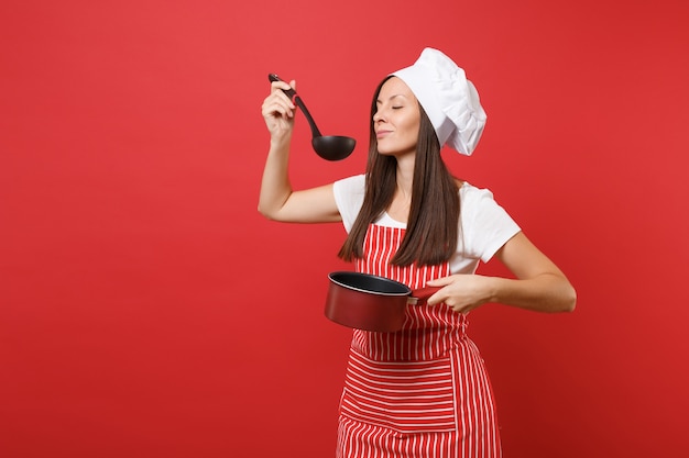 Hausfrau Köchin oder Bäckerin in gestreifter Schürze weißes T-Shirt Haube Kochmütze isoliert auf rotem Wandhintergrund. Frau halten Verkostung leerer Eintopf schwarze Suppenkelle Schöpflöffel. Mock-up-Kopierraumkonzept