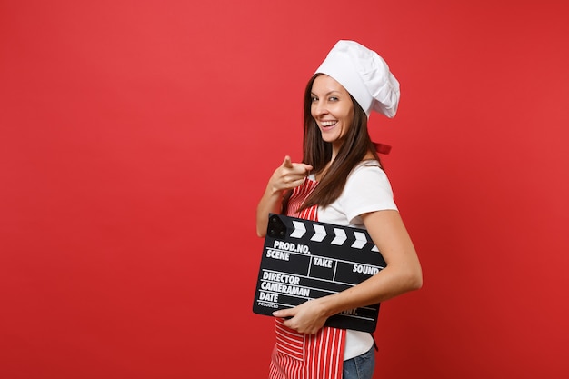 Hausfrau Köchin oder Bäckerin in gestreifter Schürze, weißes T-Shirt, Haubenköche isoliert auf rotem Wandhintergrund. Frau, die klassischen schwarzen Film hält, der Filmklappe macht. Mock-up-Kopierraumkonzept.