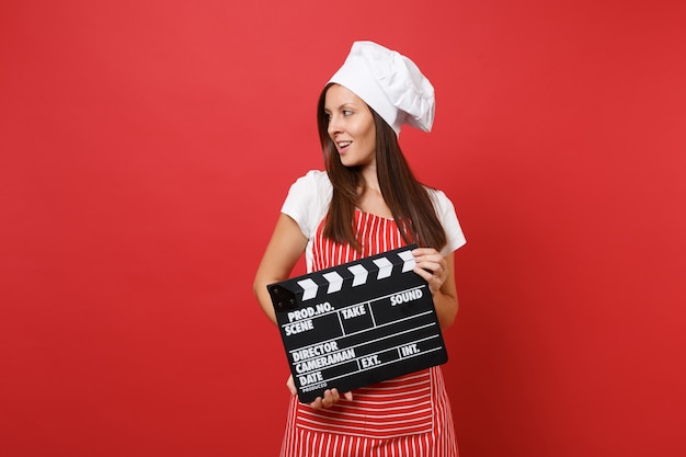 Hausfrau köchin oder bäckerin in gestreifter schürze, weißes t-shirt, haubenköche isoliert auf rotem wandhintergrund. frau, die klassischen schwarzen film hält, der filmklappe macht. mock-up-kopierraumkonzept.