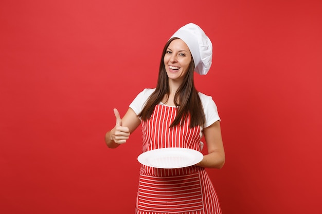 Hausfrau Köchin oder Bäckerin in gestreifter Schürze, weißes T-Shirt, Haubenköche isoliert auf rotem Wandhintergrund. Frau hält leere leere runde Platte mit Platz für Essen. Mock-up-Kopierraumkonzept