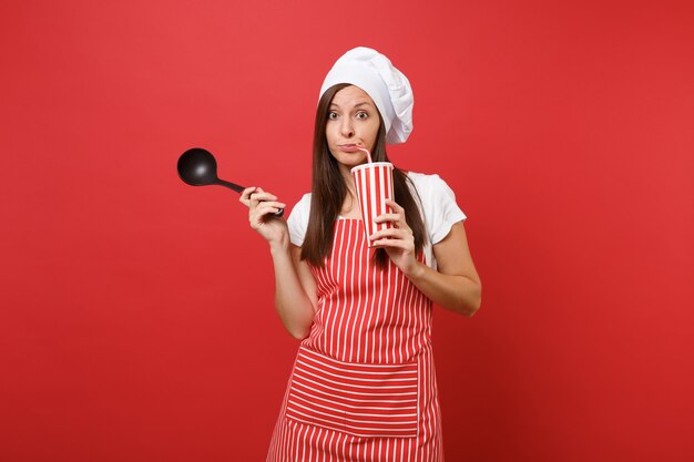 Hausfrau Köchin oder Bäckerin in gestreifter Schürze, weißes T-Shirt, Haubenköche isoliert auf rotem Wandhintergrund. Frau hält Suppe schwarze Schöpflöffel, Plastikbecher Cola. Mock-up-Kopierraumkonzept.