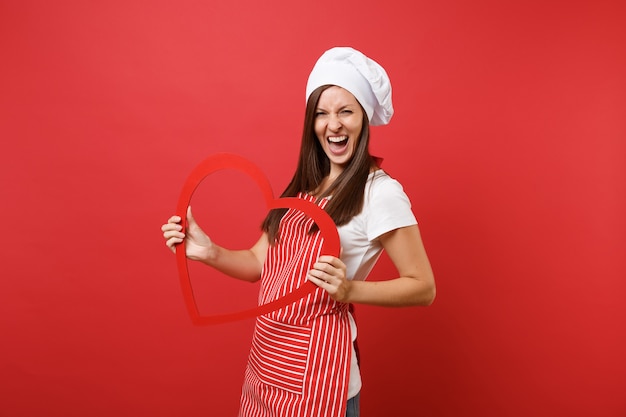Hausfrau Köchin oder Bäckerin in gestreifter Schürze, weißes T-Shirt, Haubenköche isoliert auf rotem Wandhintergrund. Lächelnde Haushälterinfrau, die hölzernes rotes Herz hält. Mock-up-Kopierraumkonzept.
