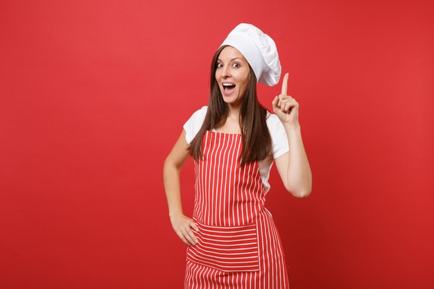 Hausfrau Köchin oder Bäckerin in gestreifter Schürze, weißes T-Shirt, Haubenköche isoliert auf rotem Wandhintergrund. Schöne Haushälterin, die mit dem Zeigefinger nach oben zeigt. Mock-up-Kopierraumkonzept.