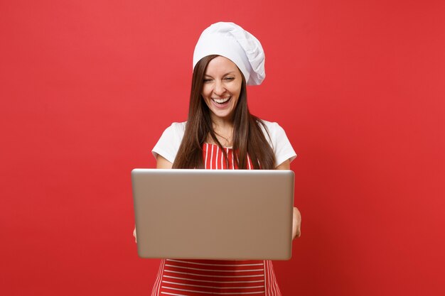 Hausfrau Köchin oder Bäckerin in gestreifter Schürze, weißes T-Shirt, Haubenköche isoliert auf rotem Wandhintergrund. Spaß Haushälterin Frau auf der Suche nach Rezept im Laptop-PC. Mock-up-Kopierraumkonzept.