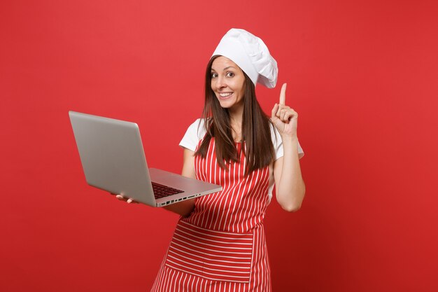 Hausfrau Köchin oder Bäckerin in gestreifter Schürze, weißes T-Shirt, Haubenköche isoliert auf rotem Wandhintergrund. Spaß Haushälterin Frau auf der Suche nach Rezept im Laptop-PC. Mock-up-Kopierraumkonzept.