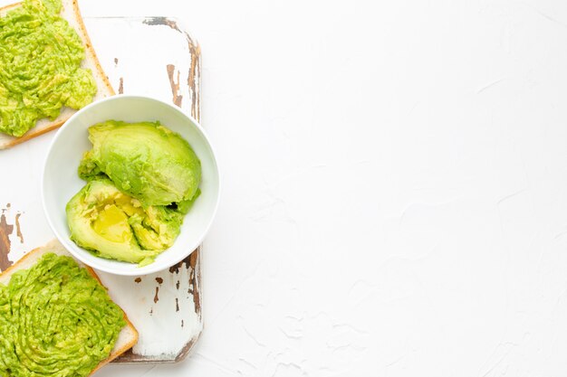 Hausgemachte Avocado-Toasts und zertrümmerte frische reife Avocados in Schüssel auf rustikalem Holzbrett auf weißem Hintergrund. Avocado-Smash-Sandwiches, gesunder Snack für die Ernährung, sauberes Essen. Ansicht von oben, Textfreiraum