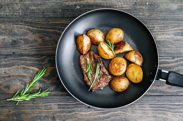 Hausgemachte Bratkartoffeln mit Rindersteak mit Rosmarin in einer Pfanne auf einem Holztisch