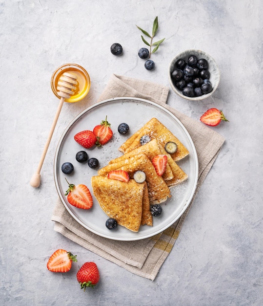 Hausgemachte dünne Pfannkuchen oder Crêpes mit frischen Heidelbeeren und Erdbeeren und Honig auf einem Teller auf blauem Hintergrund Draufsicht