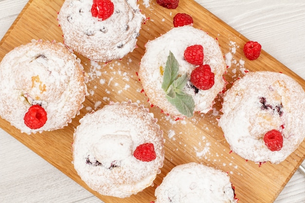 Hausgemachte Fruchtmuffins mit Puderzucker und frischen Himbeeren auf Holzbrett bestreut. Ansicht von oben