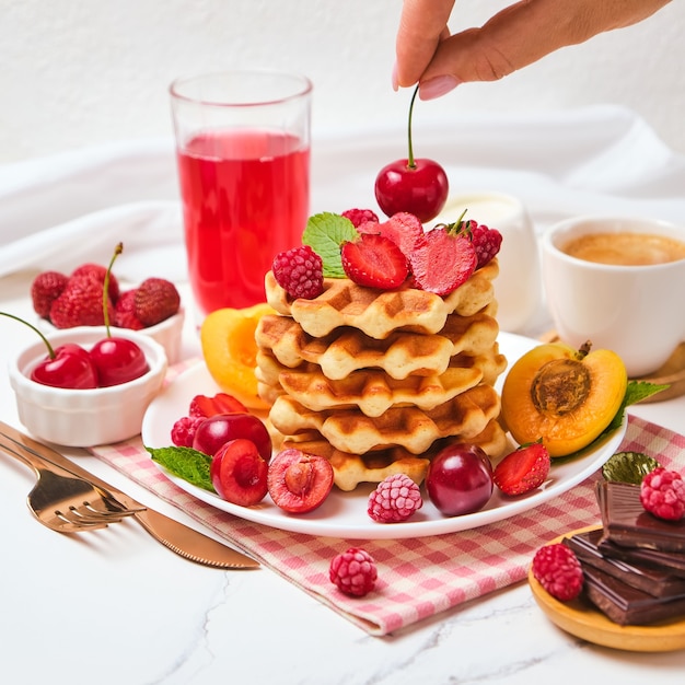 Hausgemachte gebackene traditionelle belgische Waffeln mit Beerenfrüchten und Tasse Kaffee, selektiver Fokus. weibliche Hand legt Kirschen auf Waffeln