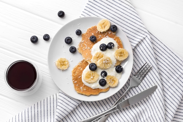 Hausgemachte Haferflockenpfannkuchen mit Joghurt, frischen Blaubeeren und Banane am weißen Holztisch.