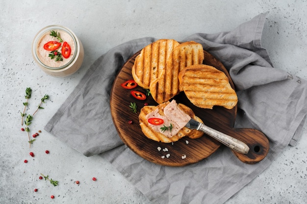 Hausgemachte Hühnchenpastete aus Leber mit rotem Pfeffer, Thymianzweigen und gerösteten Brotscheiben auf altem Holzbrett auf grauem Beton oder Stein.