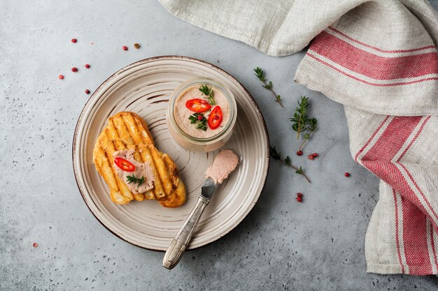 Hausgemachte Hühnchenpastete aus Leber mit rotem Pfeffer, Thymianzweigen und gerösteten Brotscheiben auf altem Holzbrett auf grauem Beton oder Stein.