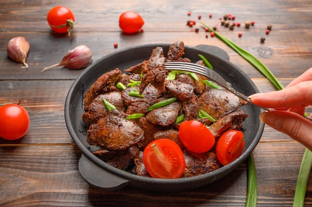 Hausgemachte Hühnerleber gebraten mit Sojasauce, Tomaten, Zwiebeln und Gewürzen auf Holztisch. Frauenhand hält eine Gabel mit einem gestochenen Stück