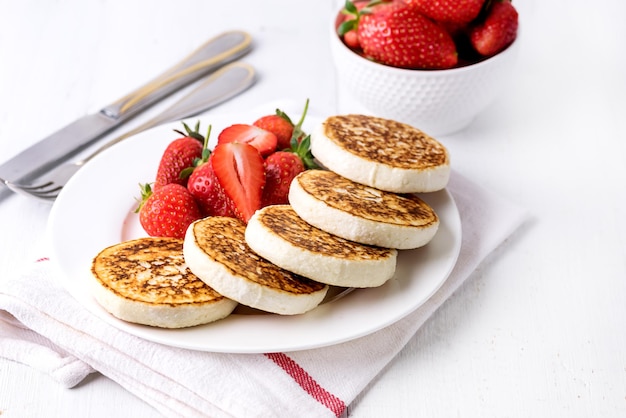 Hausgemachte Hüttenkäse-Pfannkuchen mit reifen Erdbeeren auf weißem Teller auf weißem Holzhintergrund