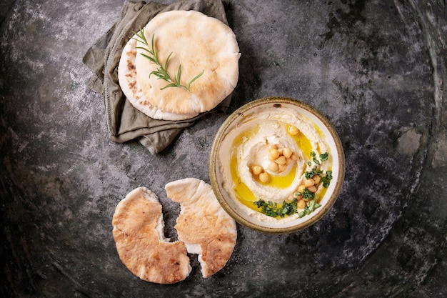 Hausgemachte Hummusschale, dekoriert mit gekochten Kichererbsen, Kräutern, Pita und Olivenöl über einem rustikalen Metallhintergrund. Draufsicht.