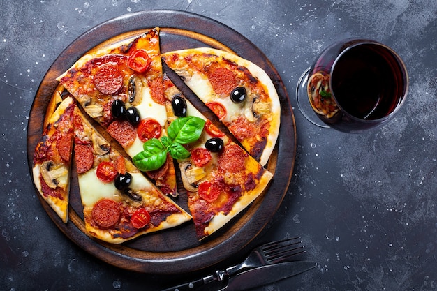 Hausgemachte italienische Pizza mit Mozzarella, Peperoni-Würstchen, Oliven und Basilikum und einem Glas Rotwein auf der Tischplattenansicht.