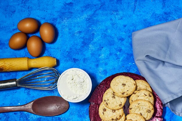 Hausgemachte Kekse im Teller mit Eiern und Schneebesen und Eiermehl auf blauem Grunge-Hintergrund