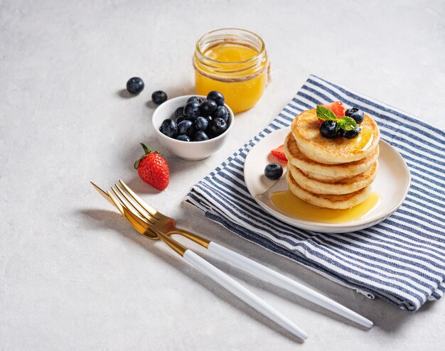 Hausgemachte klassische amerikanische Pfannkuchen in einem Stapel mit frischen Beeren und Honig auf einer gestreiften Serviette auf einem hellgrauen Texturhintergrund. Das Konzept eines gesunden und diätetischen Frühstücks. Platz für Text.