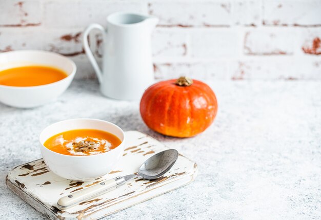 Hausgemachte Kürbiscremesuppe serviert in weißer Keramikschale auf weißem Tisch mit Löffel verziert mit ganzem Kürbis, Winkelansicht, selektiver Fokus. Herbstliches gemütliches Komfortessen, Hintergrundraum für Text