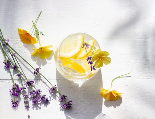 Hausgemachte Limonade mit Zitrone und Eiswürfeln in einem Glas auf dem weißen Holztisch mit gelben Blüten und Lavendel