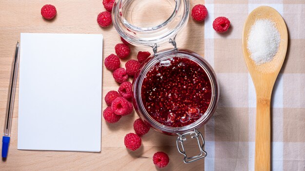 Hausgemachte Marmelade im Glas und gestreute Himbeeren auf dem Tisch ein Löffel mit Zucker und Rezeptpapier