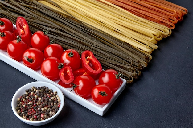 Hausgemachte Nudeln in verschiedenen Farben aus natürlichen Kräuterzutaten eine längliche Schüssel mit Tomaten und rotem Paprika und eine Schüssel mit bunten Paprika auf einem schwarzen Betontisch