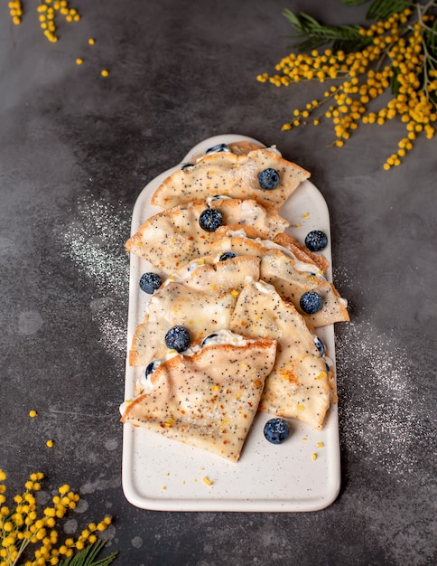 Hausgemachte Pfannkuchen, Crepes mit frischen Blaubeeren, Frischkäse und Puderzucker. Leckeres Dessert. Dunkle Oberfläche mit Frühlingsblumen. vertikales Bild.