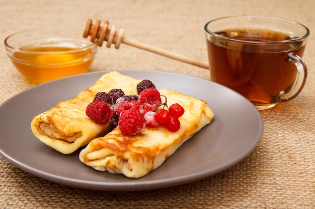 Hausgemachte Pfannkuchen gefüllt mit Hüttenkäse und belegt mit gefrorenen Himbeeren und Brombeeren auf Teller, Tasse Tee und Honig in Glasschüssel mit Holzlöffel im Hintergrund