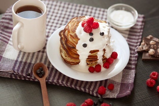 hausgemachte Pfannkuchen mit saurer Sahne auf weißem Teller mit Beeren, frischen Blaubeeren und Himbeeren, Tasse Tee oder Kaffee, Löffel, Chocholatstücke auf kariertem Baumwolltuch.