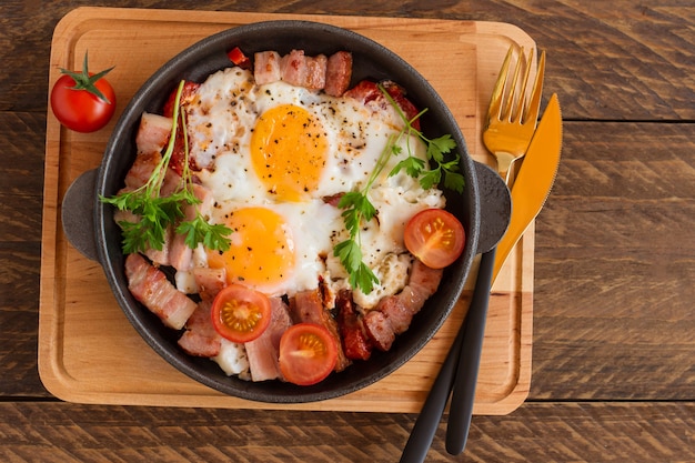 Hausgemachte Spiegeleier mit Speck und Tomaten in einer Pfanne auf Holzhintergrund. Klassisches Frühstück. Ansicht von oben.