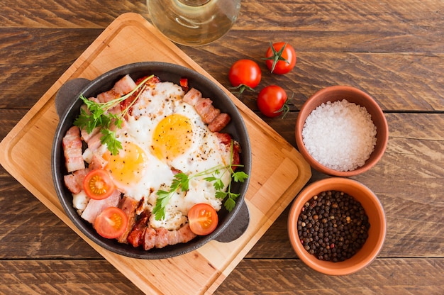 Hausgemachte Spiegeleier mit Speck und Tomaten in einer Pfanne auf Holzhintergrund. Klassisches Frühstück. Ansicht von oben.