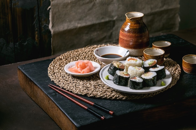 Hausgemachte Sushi-Rollen mit Lachs, japanischem Omelett, Avocado, Ingwer und Sojasauce mit Stäbchen auf Strohserviette. Schwarzer Holztisch. Sake-Set aus Keramik. Abendessen im japanischen Stil