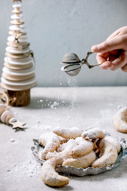 Hausgemachte traditionelle weihnachts-mürbeteig-vanille-halbmonde mit puderzucker, der vom sieb besprüht wird. auf keramikplatte mit hölzernen weihnachtsdekorationen über hellgrauer oberfläche.