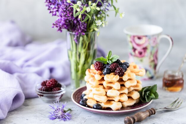 Hausgemachte Waffeln mit Beeren und Honig, eine Tasse Kaffee auf dem Tisch mit einem Strauß Flieder.