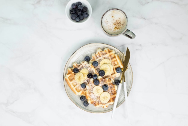 Hausgemachte Wiener Waffeln mit Heidelbeeren und Banane und Tasse Kaffee auf Marmorhintergrund