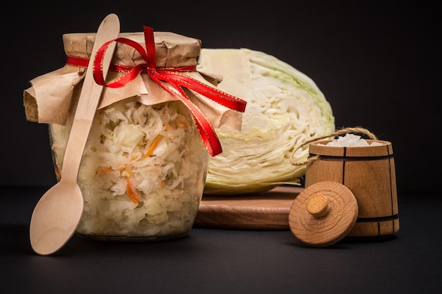 Hausgemachter fermentierter Kohl im Glas auf schwarzem Hintergrund. Frischer Kohlkopf und Salz im Hintergrund. Veganer Salat. Das Gericht ist reich an Vitamin U. Nahrung für eine gute Gesundheit.