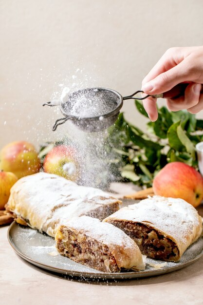 Hausgemachter geschnittener klassischer Apfelstrudel, bestreut mit Puderzucker aus dem Sieb in der Hand, auf Keramikplatte mit frischen Äpfeln, grünen Blättern und Zimtstangen darüber.