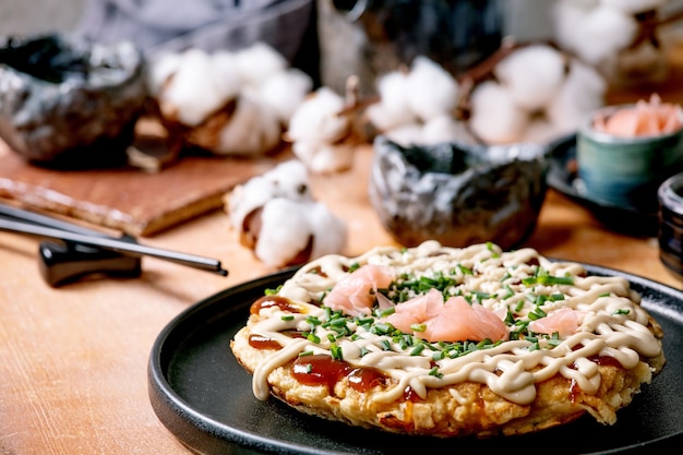 Hausgemachter japanischer Fast-Food-Okonomiyaki-Kohlpfannkuchen mit Zwiebeln, eingelegtem Ingwer, Mayo-Sauce auf schwarzem Keramikteller. Essstäbchen, Teekanne, Baumwolle, Zutaten oben .. Flach legen