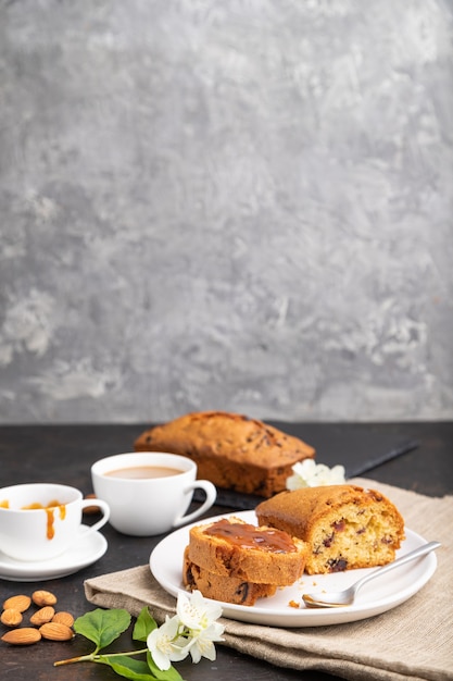 Hausgemachter Kuchen mit Rosinen, Mandeln, weichem Karamell und einer Tasse Kaffee auf einer schwarzen Betonoberfläche und Leinentextil