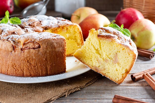 Hausgemachter runder Biskuitkuchen oder Chiffonkuchen mit Äpfeln auf weißem Teller so weich und lecker mit Zutaten: Eier, Mehl, Milch auf dem Tisch. Hausgemachtes Bäckereikonzept für Hintergrund und Tapete.