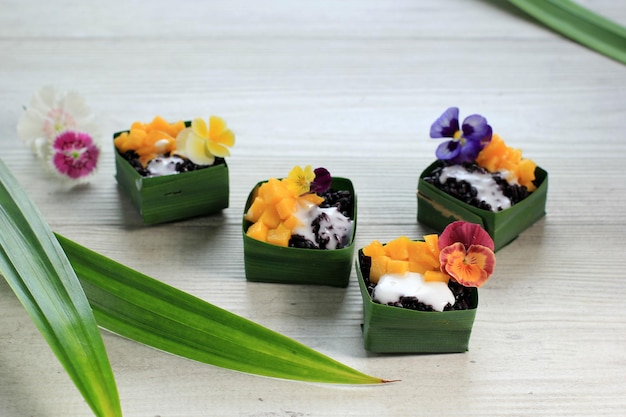 Hausgemachter schwarzer Kokos-Klebreis mit Mango in Pandan-Blätter-Becher, selektiver Fokus mit Stiefmütterchen-Edibel-Blumen-Topping