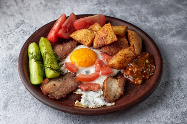 Hausgemachtes frühstück auf einem teller, gesalzene gurken, bratkartoffeln, spiegelei, speck, tomaten und hausgemachte sauce