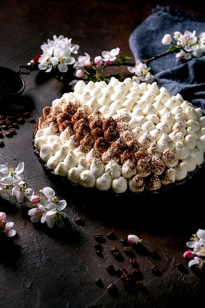 Hausgemachtes glutenfreies Tiramisu traditionelles italienisches Dessert mit Kakaopulver bestreut mit blühendem Apfelbaum, blauer Textilserviette und Kaffeebohnen über dunkler Texturoberfläche.