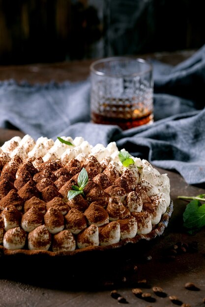 Hausgemachtes glutenfreies Tiramisu traditionelles italienisches Dessert mit Kakaopulver bestreut mit Minzblättern, Glas Whisky, blauer Textilserviette und Kaffeebohnen. Dunkle Oberfläche. Nahansicht