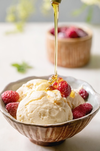 Hausgemachtes Karamell-Vanille-Eis mit fließendem Sirup, gefrorenen Himbeeren in der Keramikschale, die auf weißem Marmortisch mit Blumen dahinter steht